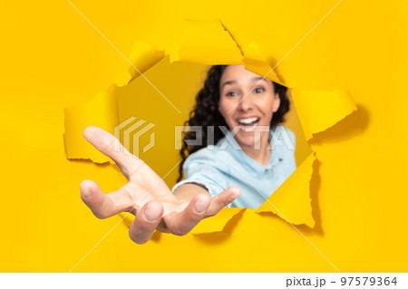 Excited woman stretching open palm through hole in torn yellow paper, taking something, holding invisible object 97579364