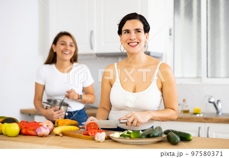 Cheerful young woman with female roommate cooking in home kitchen 97580371