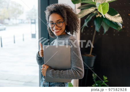 Smiling afro american woman holding laptop and looking at camera while standing near window in cafe 97582928