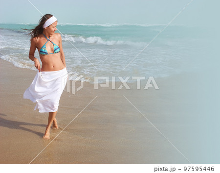 Beautiful happy girl walks barefoot, on sandy beach, along ocean, wind is blowing her hair and white skirt. Concept of travel, relaxation, happiness and freedom, copy space 97595446