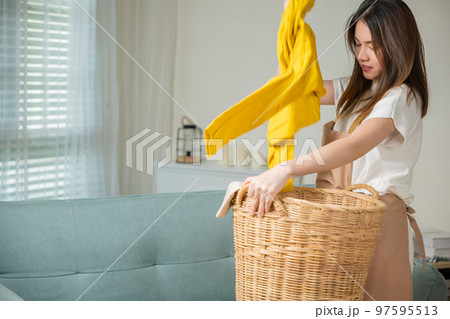 Asian housewife woman holding basket with heap of different clothes on sofa in living room, Beautiful housekeeper doing housework holds wooden basket of clean clothes to washing at home 97595513