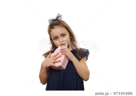 Portrait of a little girl in an elegant navy dress, isolated on white background. The child holds a pink heart-shaped box with a cute gift. The concept of celebration, giving and receiving presents Portrait of a little girl in an elegant navy dress, isolated on white background. The child holds a pink heart-shaped box with a cute gift. The concept of celebration, giving and receiving presents 97595986