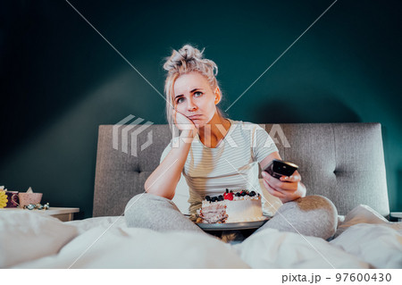 Bored, sad woman in home clothes eating cake and using the remote control to change the channel while watching television in bed at night. Overeating, Jamming of negative emotions. Selective focus Bored, sad woman in home clothes eating cake and using the remote control to change the channel while watching television in bed at night. Overeating, Jamming of negative emotions. Selective focus 97600430