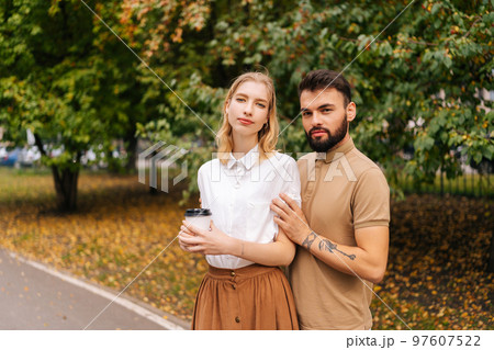 Portrait of happy young couple in love standing holding hands on city park looking at camera. Handsome bearded man with tattoo and cute blonde woman walking drinking coffee enjoying time together. 97607522