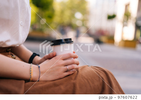 Close up side view cropped shot of unrecognizable elegant lady in casual clothes holding in hands paper cup with takeaway coffee sitting on bench on city street, in summer day, blurred background. 97607622