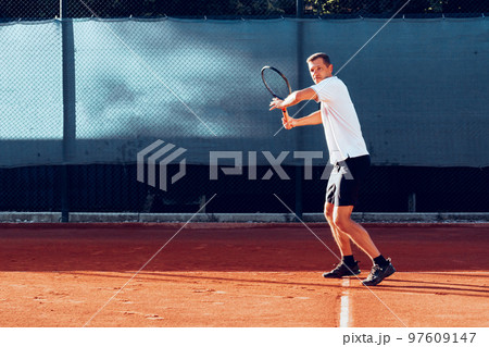 Young man plays tennis outdoors on tennis court in the morning Young man plays tennis outdoors on tennis court in the morning 97609147