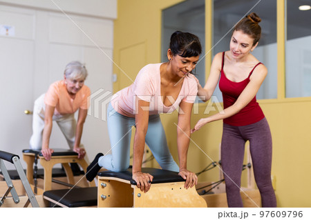 Young woman pilates instructor helping Hispanic woman exercising on combo chair Young woman pilates instructor helping Hispanic woman exercising on combo chair 97609796
