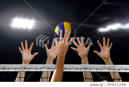Close up of Volleyball spiking and hand blocking over the net under bright spotlights Close up of Volleyball spiking and hand blocking over the net under bright spotlights 97611149
