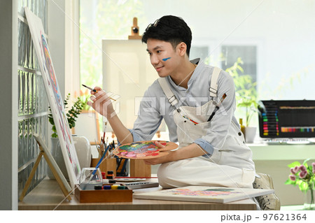 Happy asian male artist holding paintbrushes and palette painting on canvas at art studio. Education, hobby, art concept Happy asian male artist holding paintbrushes and palette painting on canvas at art studio. Education, hobby, art concept 97621364
