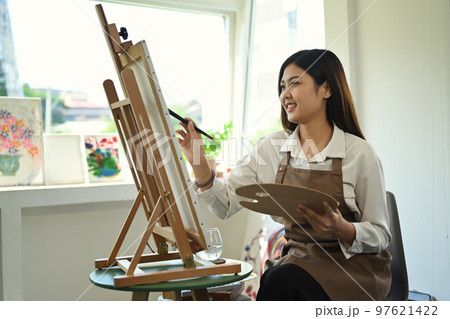 Pleasant female artist in apron with palette and brush painting on canvas at art studio. Education, hobby, art concept 97621422