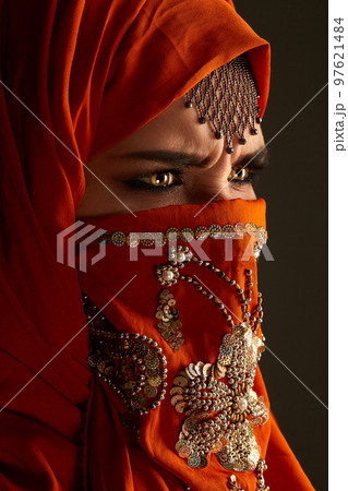 Studio shot of a young charming woman wearing the terracotta hijab decorated with sequins and jewelry. Arabic style. 97621484