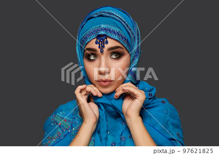 Studio shot of a young beautiful woman wearing the blue hijab decorated with sequins and jewelry. Arabic style. 97621825