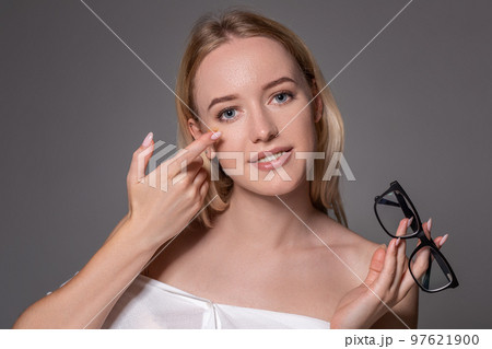 Young blonde woman holding contact lens on finger in front of her face and holding in her other hand a black glasses on gray background. 97621900