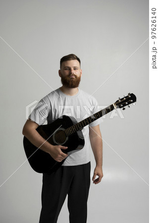 Male musician playing on acoustic guitar. Portraite of bearded handsome musician holding a guitar in a white room. Guitarist, artist, composer, rocker, star. 97626140