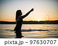 Beautiful Russian girl in a long black swimsuit swims outside the city on the lake in the rays of sunset or dawn. Splashing water on the background of the sun. Happy girl. 97630707