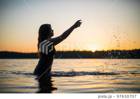 Beautiful Russian girl in a long black swimsuit swims outside the city on the lake in the rays of sunset or dawn. Splashing water on the background of the sun. Happy girl. Beautiful Russian girl in a long black swimsuit swims outside the city on the lake in the rays of sunset or dawn. Splashing water on the background of the sun. Happy girl. 97630707