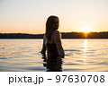 Beautiful Russian girl in a long black swimsuit swims outside the city on the lake in the rays of sunset or dawn. Splashing water on the background of the sun. Happy girl. 97630708