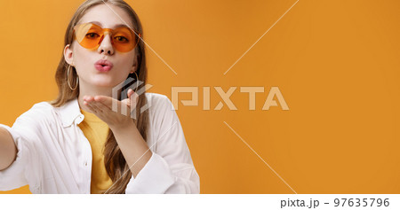 Stylish popular girl sending air kiss at camera taking selfie or recording vlog working as freelance travel advisor taking pictures against orange background tender and feminine pulling hand forward 97635796