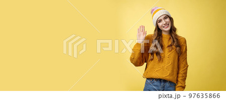 Friendly charming smiling young woman tilting head outgoing gaze waving hand hello hi gesture greeting you hey what up, standing carefree joyfully looking camera, yellow background 97635866