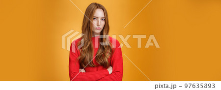 Suspicious intense and defensive ginger girl standing in passive-aggressive pose pouting and frowning looking with disbelief and disdain at camera, offended against orange background 97635893