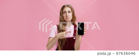 Indoor shot of gloomy displeased and disappointed cute young girl holding smartphone pointing at cellphone screen making upset grimace as if being bothered and dissatisfied against pink wall Indoor shot of gloomy displeased and disappointed cute young girl holding smartphone pointing at cellphone screen making upset grimace as if being bothered and dissatisfied against pink wall 97636046