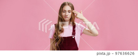 Portrait of bothered uneasy cute european female with long natural fair hair in trendy corduroy overalls smirking from irritation and annoyance showing finger fun gesture as if shooting in head Portrait of bothered uneasy cute european female with long natural fair hair in trendy corduroy overalls smirking from irritation and annoyance showing finger fun gesture as if shooting in head 97636626