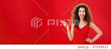 Girl suggesting ask her boyfriend. Charming elegant and stylish young female with curly hairstyle in red evening dress holding hand on hip and pointing left while showing friend where buy clothes Girl suggesting ask her boyfriend. Charming elegant and stylish young female with curly hairstyle in red evening dress holding hand on hip and pointing left while showing friend where buy clothes 97636635