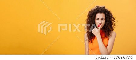 Waist-up shot of serious determined and focused displeased woman with curly hairstyle frowning biting thumb looking dangerous and angry standing daring and rebellious over orange background 97636770