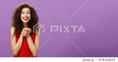 Woman happy receive award being touched and delighted holding palms together near breast smiling with tender and grateful expression standing with curly hairstyle in evening red dress over purple wall Woman happy receive award being touched and delighted holding palms together near breast smiling with tender and grateful expression standing with curly hairstyle in evening red dress over purple wall 97636809