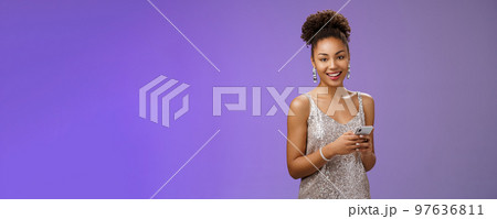 Attractive stylish confident african-american woman in silver shiny dress holding smartphone messaging scrolling internet gadget looking lucky happily camera standing blue background rejoicing 97636811