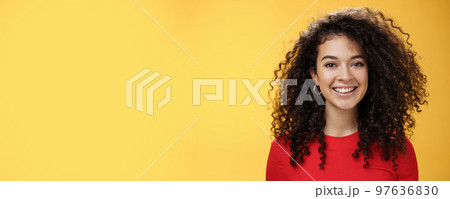 Close-up shot of pretty caucasian girl with curly hair in red dress and earrings smiling joyfully with pleased hopeful expression gazing at camera carefree, posing over yellow background 97636830