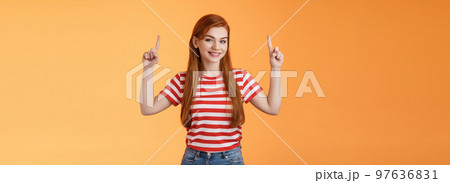 Cheerful beautiful redhead woman smiling invite try out new product, pointing up, show top copy space, grinning joyfully, rejoice introduce cool thing, stand orange background casual urban outfit 97636831