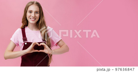 Girl loves family. Kind charming young woman in overalls with small tattoo on arm showing heart gesture over body and smiling lovely at camera expressing tender and cute attitude over pink wall 97636847