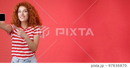 Excited happy good-looking redhead curly female show horizontal smartphone screen pointing display gadget smiling pleased proud beat friends score game standing red background 97636870