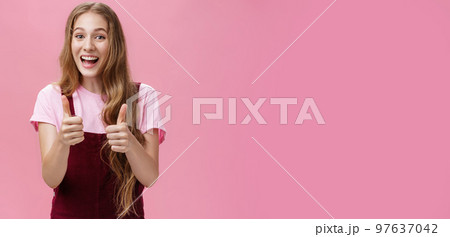 Friendly and outgoing pretty young woman in overalls with wavy natural hairstyle showing thumbs up gesture with cheer and joy smiling being supportive, liking idea, expressing positive opinion 97637042