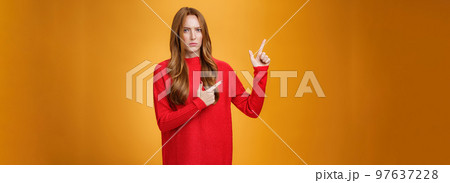 Intense suspicious and doubtful european ginger girl in warm sweater frowning hesitant and confused pointing at upper left corner asking question, looking with disbelief over orange background 97637228