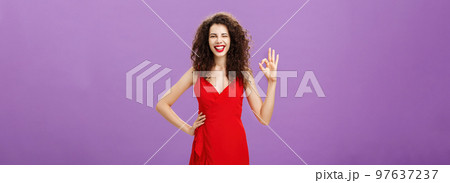 I got you covered. Happy outgoing good-looking elegant european female with curly hairstyle in red fashionable dress, make-up holding hand on waist showing okay sign in approval and smiling assuring 97637237