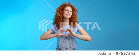 Sassy confident good-looking modern redhead curly woman raise head proud love tell girlfriend romantic heartwarming feelings present own heart smiling broadly standing blue background 97637243