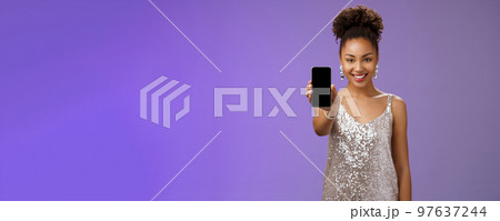 Charming attractive african-american woman in silver glittering stylish dress extend arm showing smartphone display presenting app promoting cool device standing pleased smiling blue background 97637244