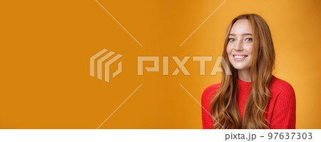 Close-up shot of happy charismatic and carefree cute redhead girl in red knittes outfit smiling joyfully as turning at camera satisfied and joyful posing delighted over orange background 97637303