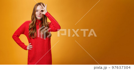 I got you on aim. Self-assured expressive and confident attractive redhead woman in red warm dress showing okay or zero sign on eye and peeking through as smiling satisfied over orange wall 97637304