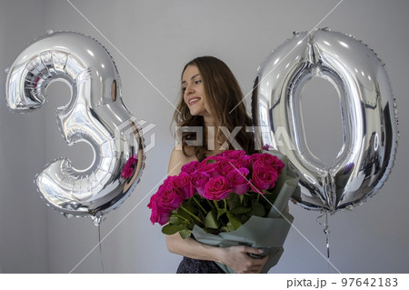 Beautiful Woman with Curls in a Dress Embraces a Big Bouquet of Flowers. Girl with Pink Roses Next to Inflatable Figures Celebrates Her Anniversary and Birthday Thirty Years. 97642183