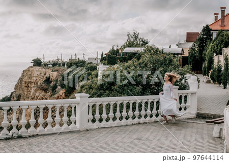 Romantic bride, blond girl in white wedding dress with open shoulders posing on open terrace with backdrop of the sea and rocks. Stylish young woman standing on terrace and looking on ocean at sunset 97644114
