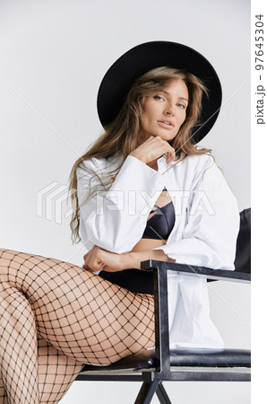 Beautiful brunette girl sits on chair and flirts, she is dressed in mesh tights, white shirt and black hat, long hair, pure white background, soft light Beautiful brunette girl sits on chair and flirts, she is dressed in mesh tights, white shirt and black hat, long hair, pure white background, soft light 97645304