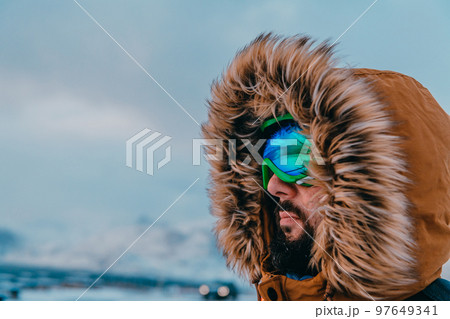 Headshot photo of a man in a cold snowy area wearing a thick brown winter jacket, snow goggles and gloves. Life in cold regions of the country. Headshot photo of a man in a cold snowy area wearing a thick brown winter jacket, snow goggles and gloves. Life in cold regions of the country. 97649341