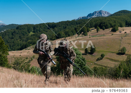 A sniper team squad of soldiers is going undercover. Sniper assistant and team leader walking and aiming in nature with yellow grass and blue sky. Tactical camouflage uniform. 97649363