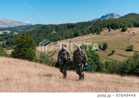 A sniper team squad of soldiers is going undercover. Sniper assistant and team leader walking and aiming in nature with yellow grass and blue sky. Tactical camouflage uniform. A sniper team squad of soldiers is going undercover. Sniper assistant and team leader walking and aiming in nature with yellow grass and blue sky. Tactical camouflage uniform. 97649449
