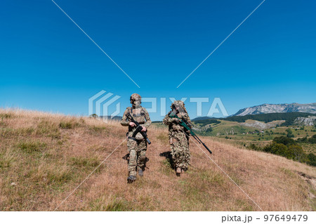 A sniper team squad of soldiers is going undercover. Sniper assistant and team leader walking and aiming in nature with yellow grass and blue sky. Tactical camouflage uniform. 97649479