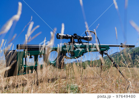 A green military sniper rifle with a scope for long distance tactical modern warfare in yellow grass blue sky A green military sniper rifle with a scope for long distance tactical modern warfare in yellow grass blue sky 97649480