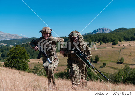 A sniper team squad of soldiers is going undercover. Sniper assistant and team leader walking and aiming in nature with yellow grass and blue sky. Tactical camouflage uniform. A sniper team squad of soldiers is going undercover. Sniper assistant and team leader walking and aiming in nature with yellow grass and blue sky. Tactical camouflage uniform. 97649514